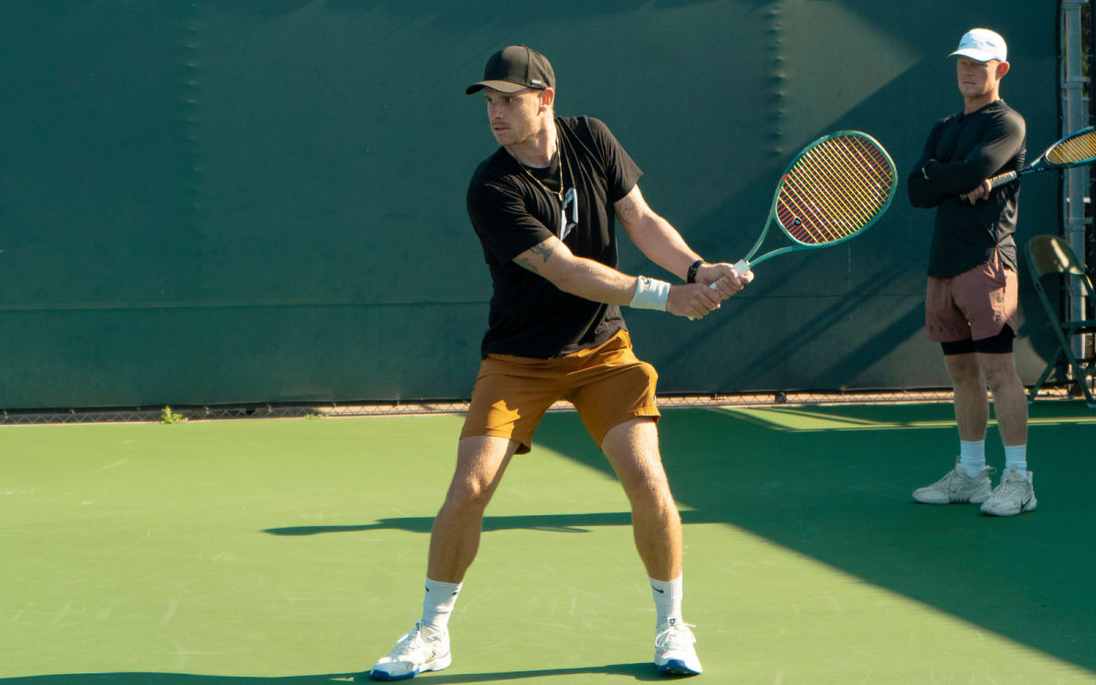 A touring professional in ready position on a practice court, a second player watching