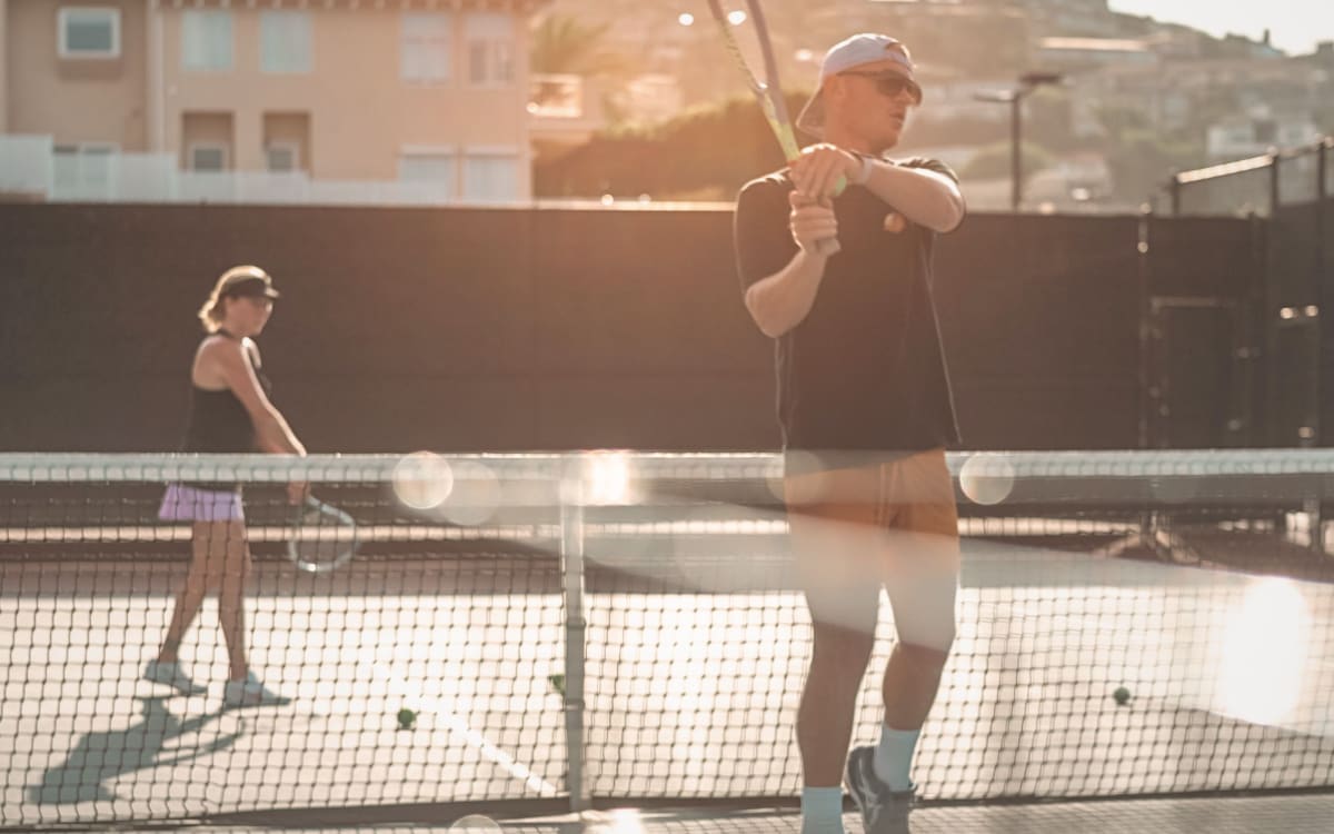 A coach at the net in golden evening light, a player rallying behind