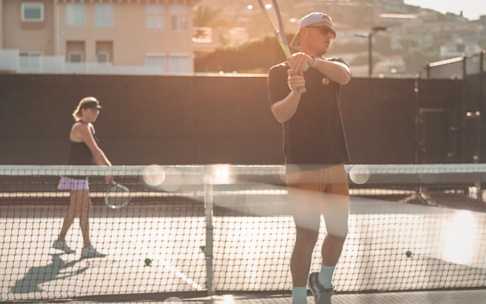 A coach at the net in golden evening light, a player rallying behind