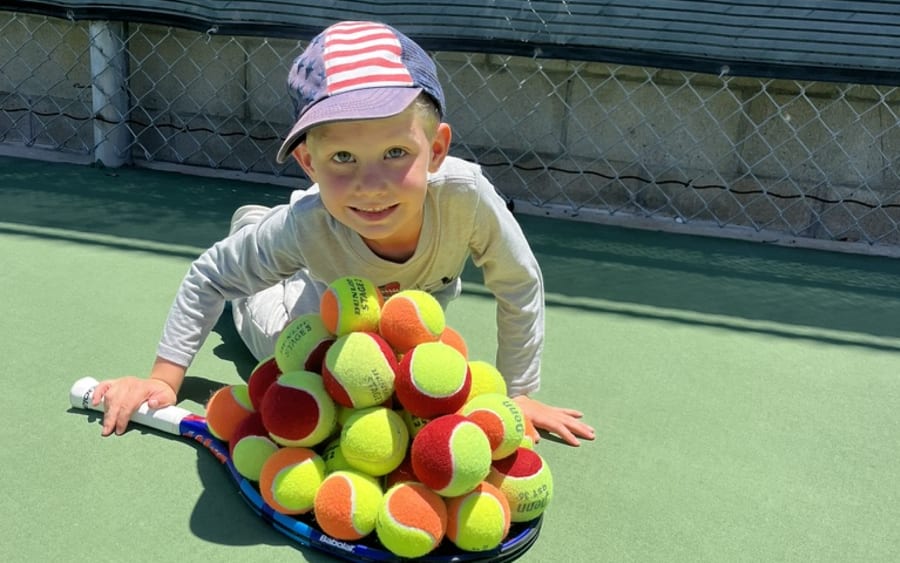 A young child on court behind a pyramid of red and orange junior balls, racquet in hand